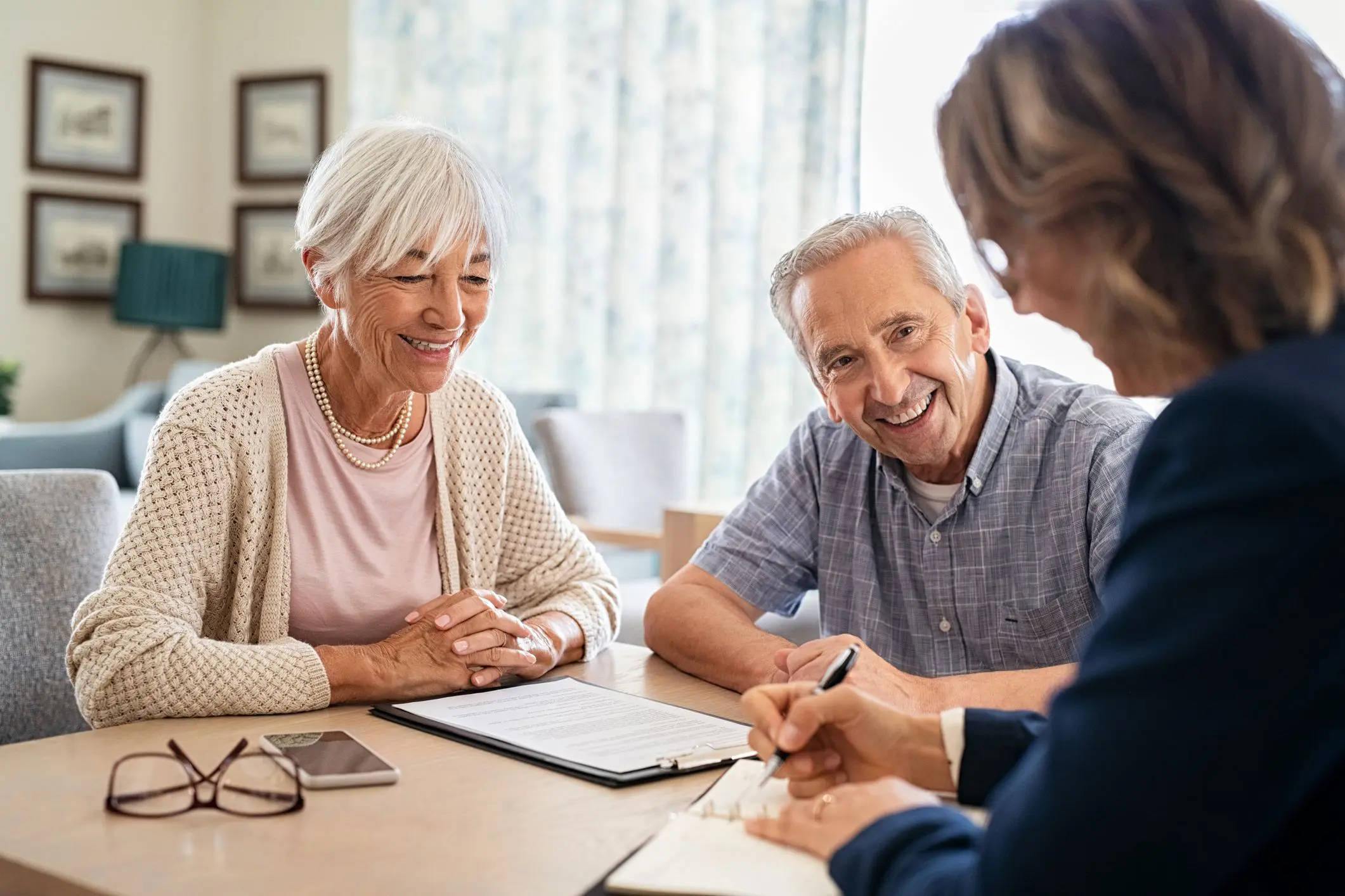 jpeg-optimizer_GettyImages-1316202262 An elderly man and woman sit at a table, smiling, as they discuss paperwork with another person.