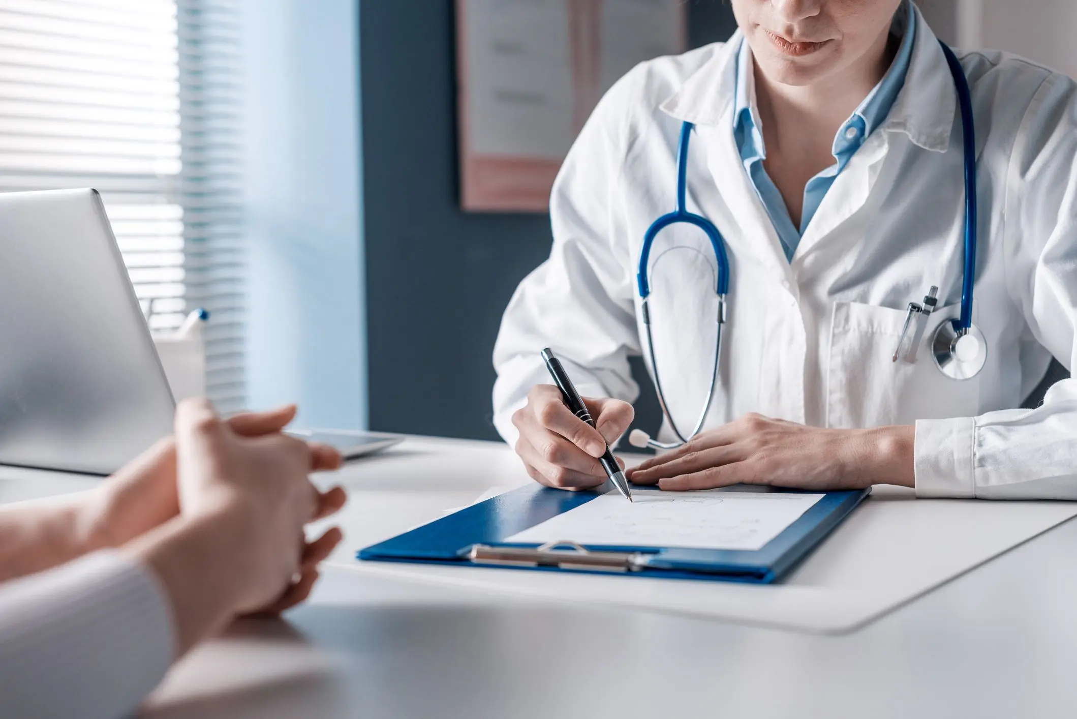 Doctor in a white coat and stethoscope writing on a clipboard at a desk while a patient sits opposite, with hands clasped.