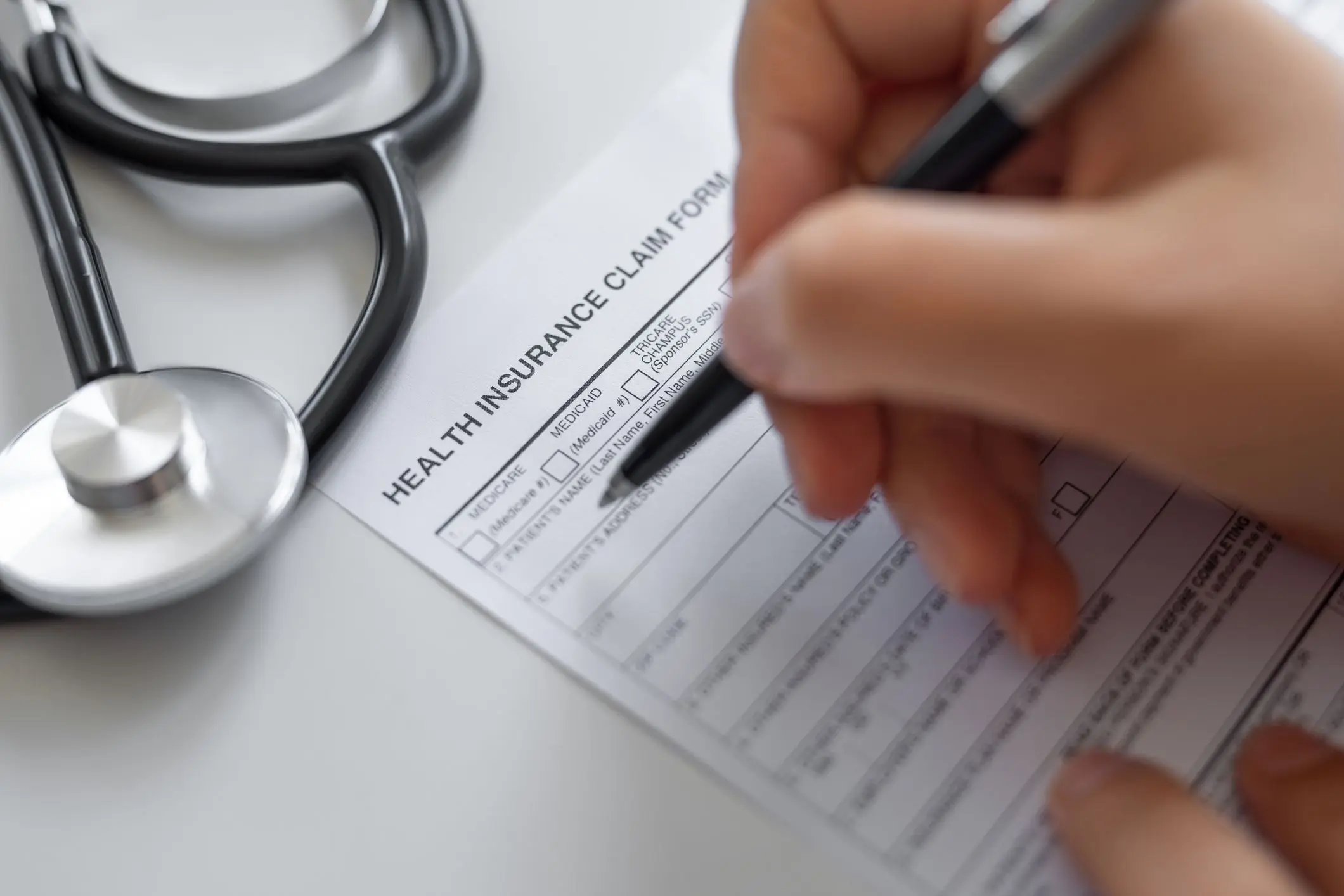 jpeg-optimizer_GettyImages-1759307341 A person fills out a health insurance claim form with a black pen. A stethoscope is visible on the table next to the form.