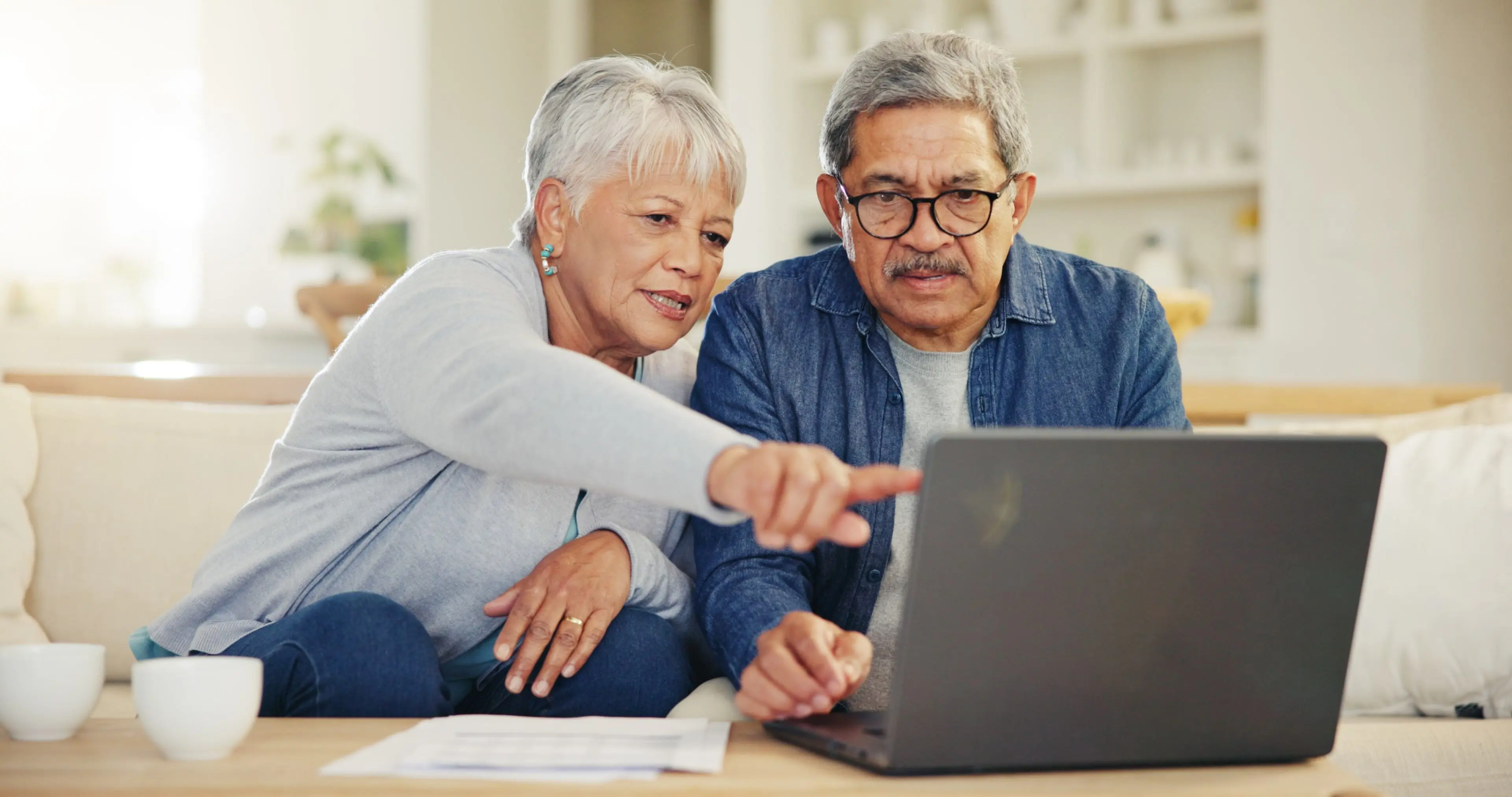 Elderly couple sitting on a couch, looking intently at a laptop screen. The woman is pointing at the screen while the man observes, wearing glasses.