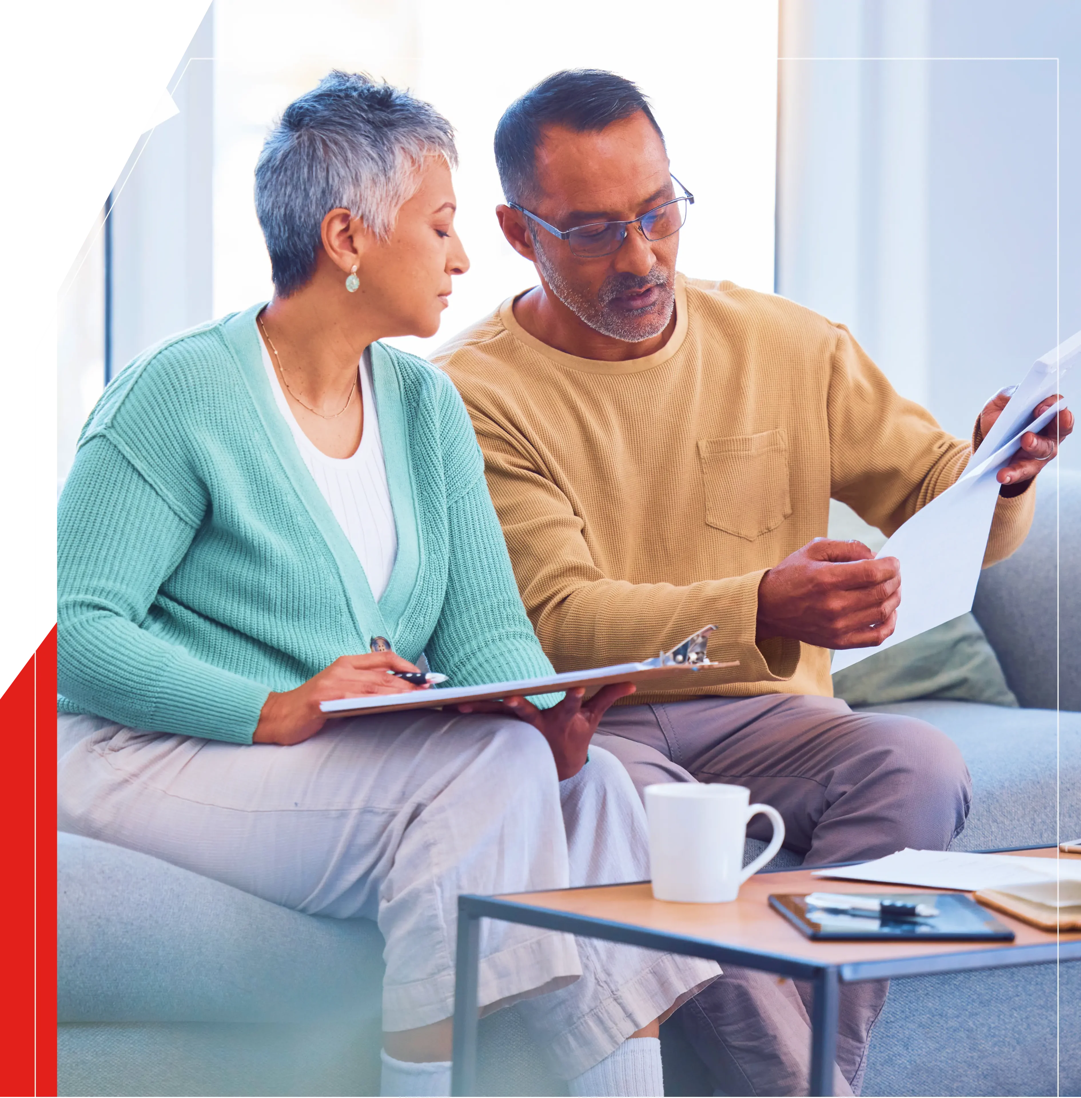 Group 1 A man and woman sit on a couch reviewing documents. The man wears glasses and a beige sweater; the woman is in a green cardigan. A coffee table with items is in front of them.